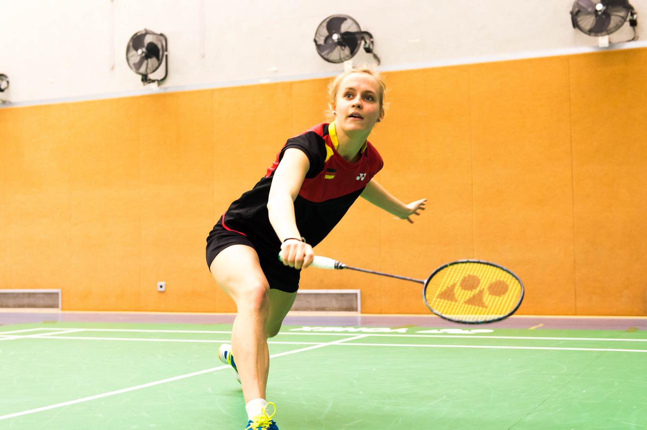 Spielerin schlägt einen Badmintonball auf einer grünen Spielfläche in einer Sporthalle mit orangefarbenen Wänden.