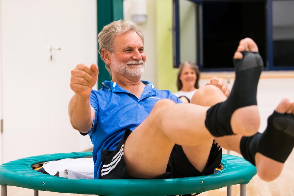 Älterer Mann macht Übungen auf einem Trampolin, lächelnd, mit einem anderen Teilnehmer im Hintergrund.
