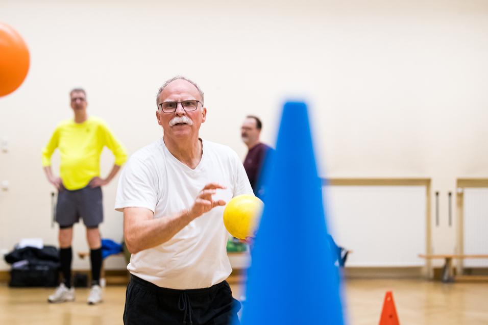 Mann mit Brille wirft einen gelben Ball in einem Sportraum, während andere Teilnehmer im Hintergrund stehen.