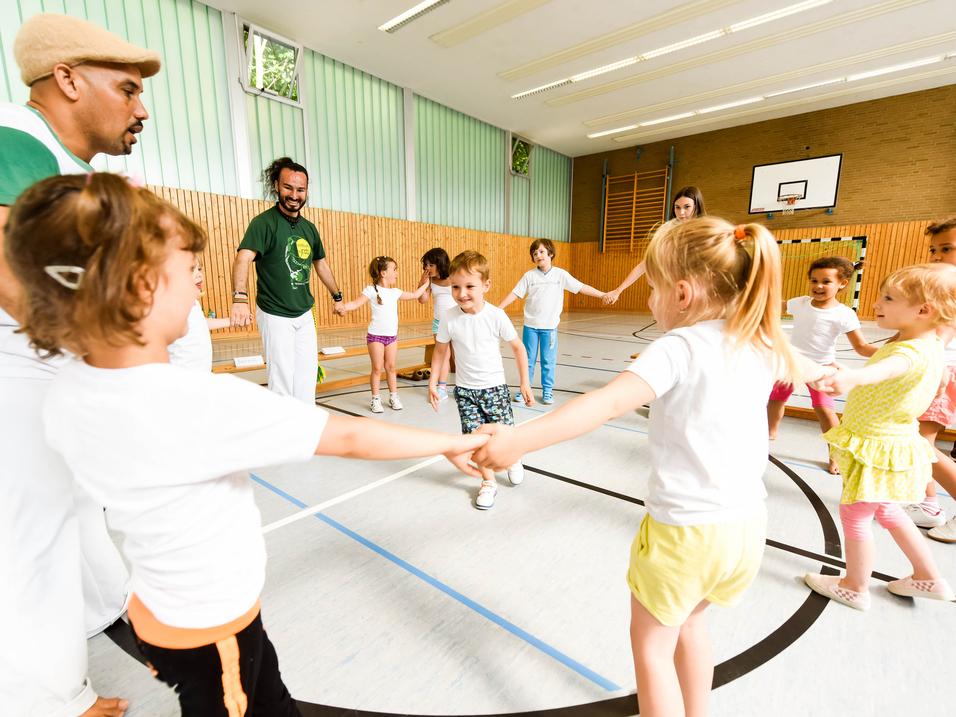Gruppe von Kindern in T-Shirts und Erwachsenen in einer Turnhalle, die gemeinsam einen Kreis bilden und spielen.