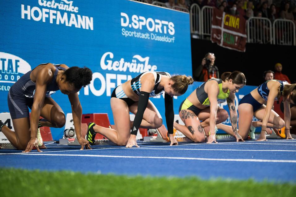 Vier Athletinnen in Startposition auf einer blauen Tartanbahn bei einem Wettkampf, mit Sponsorenbanner im Hintergrund.