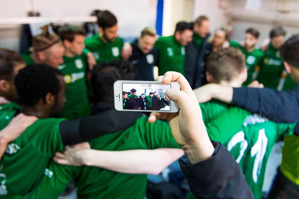 Hand hält ein Smartphone, das eine Gruppe von Spielern in grünen Trikots während einer Besprechung aufnimmt.