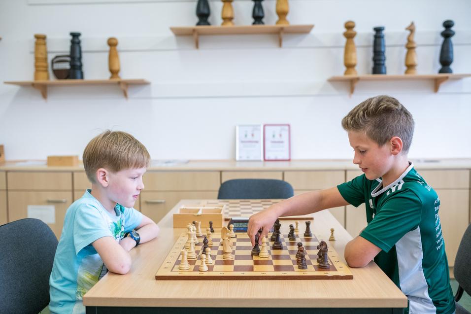 Zwei Kinder spielen Schach an einem Holztisch, mit Figuren auf dem Schachbrett und einer digitalen Uhr.