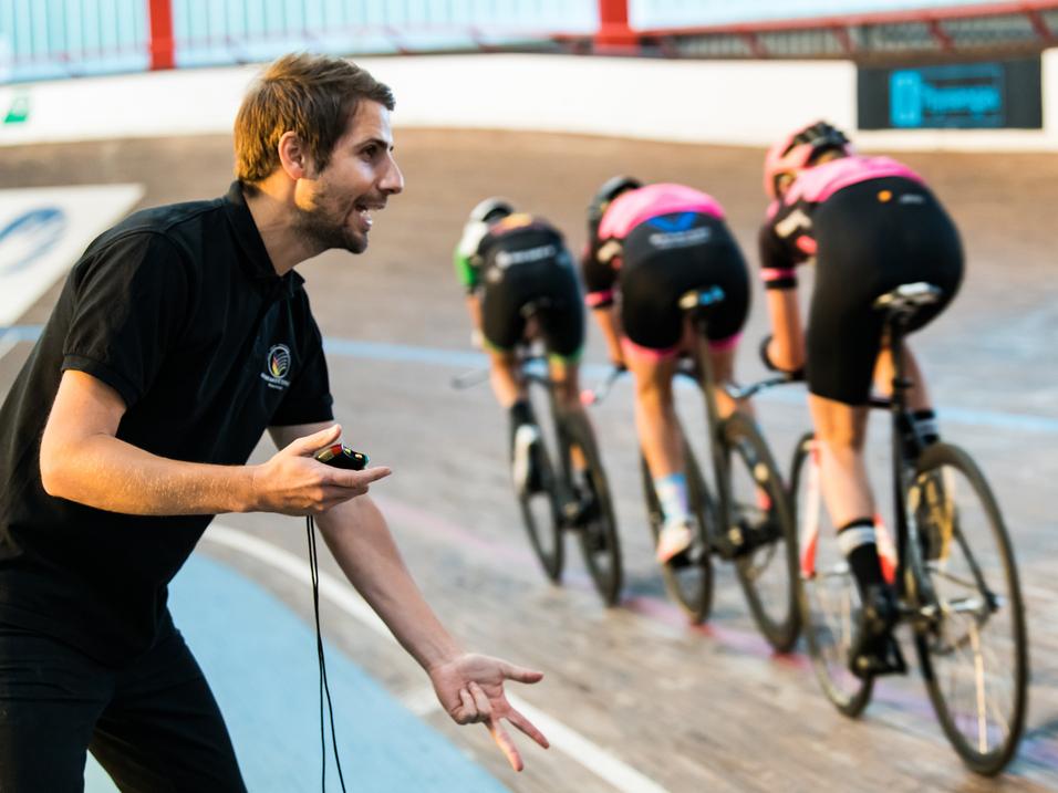 Trainer gibt Anweisungen an Gruppe von Radfahrerinnen auf einer Bahn. Vier Fahrerinnen in schwarzen und pinken Trikots.