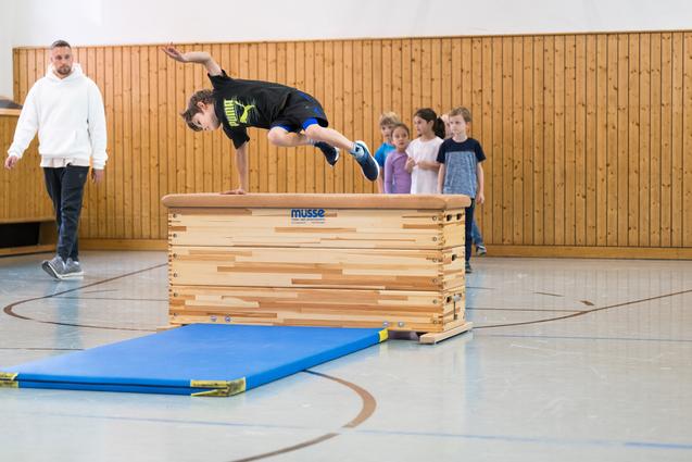 Ein Junge springt über ein Turngerät, während eine Gruppe Kinder und ein Trainer zuschauen. Sporthalle mit Holzverkleidung.